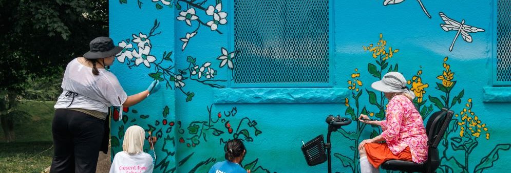Two adults and two children painting a wall mural with a blue background, white flowers and yellow flowers, and white dragonflies.
