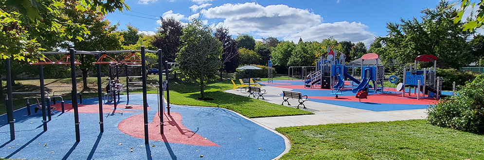A variety of outdoor fitness equipment in the foreground and a playground with swings in the background.