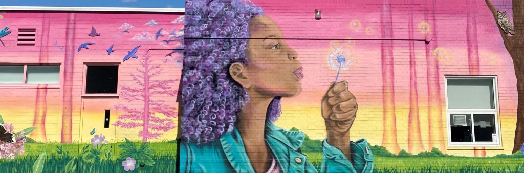Mural covering the side of a one storey building, depicts a figure in profile with purple hair set against a colourful background of magenta, pink and yellow.