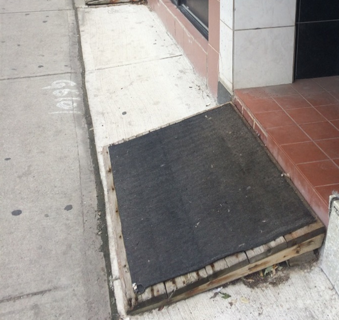 Image with the black ramp - A city sidewalk scene with a damaged, makeshift wooden ramp at a storefront entrance. The ramp is unstable, misaligned with the step, has gaps along its sides, and creates wayfinding challenges. The image illustrates unsafe, non-compliant ramp conditions.