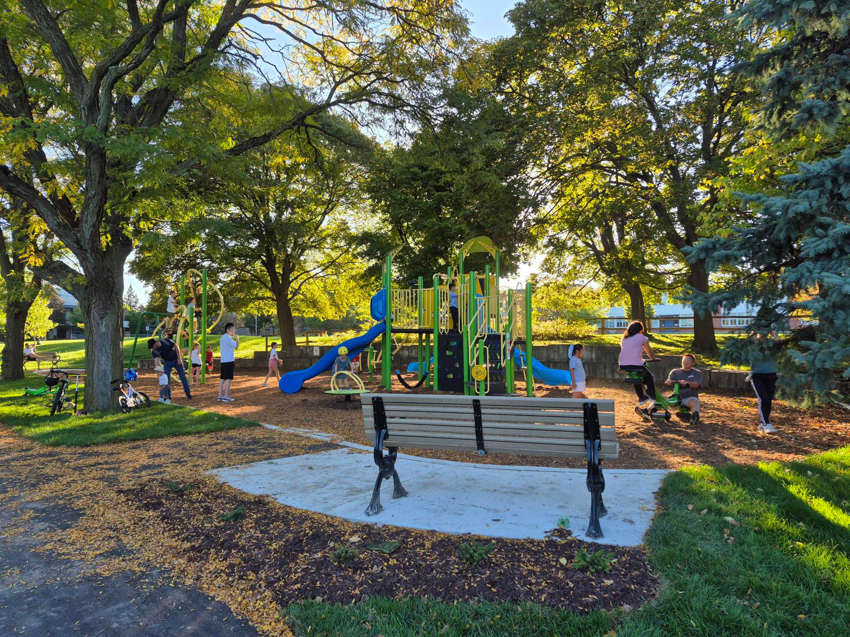 A brightly coloured playground with people using it. A bench on a paved slab is in the foreground.