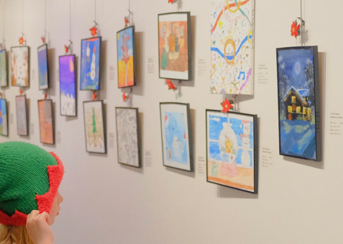 A child standing in front of artwork on a gallery wall, pointing to a painting of a human figure wearing a red and white Christmas hat.