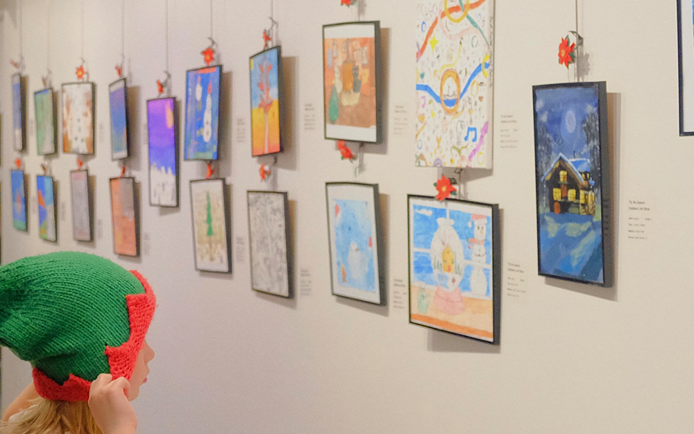 A child standing in front of artwork on a gallery wall, pointing to a painting of a human figure wearing a red and white Christmas hat.
