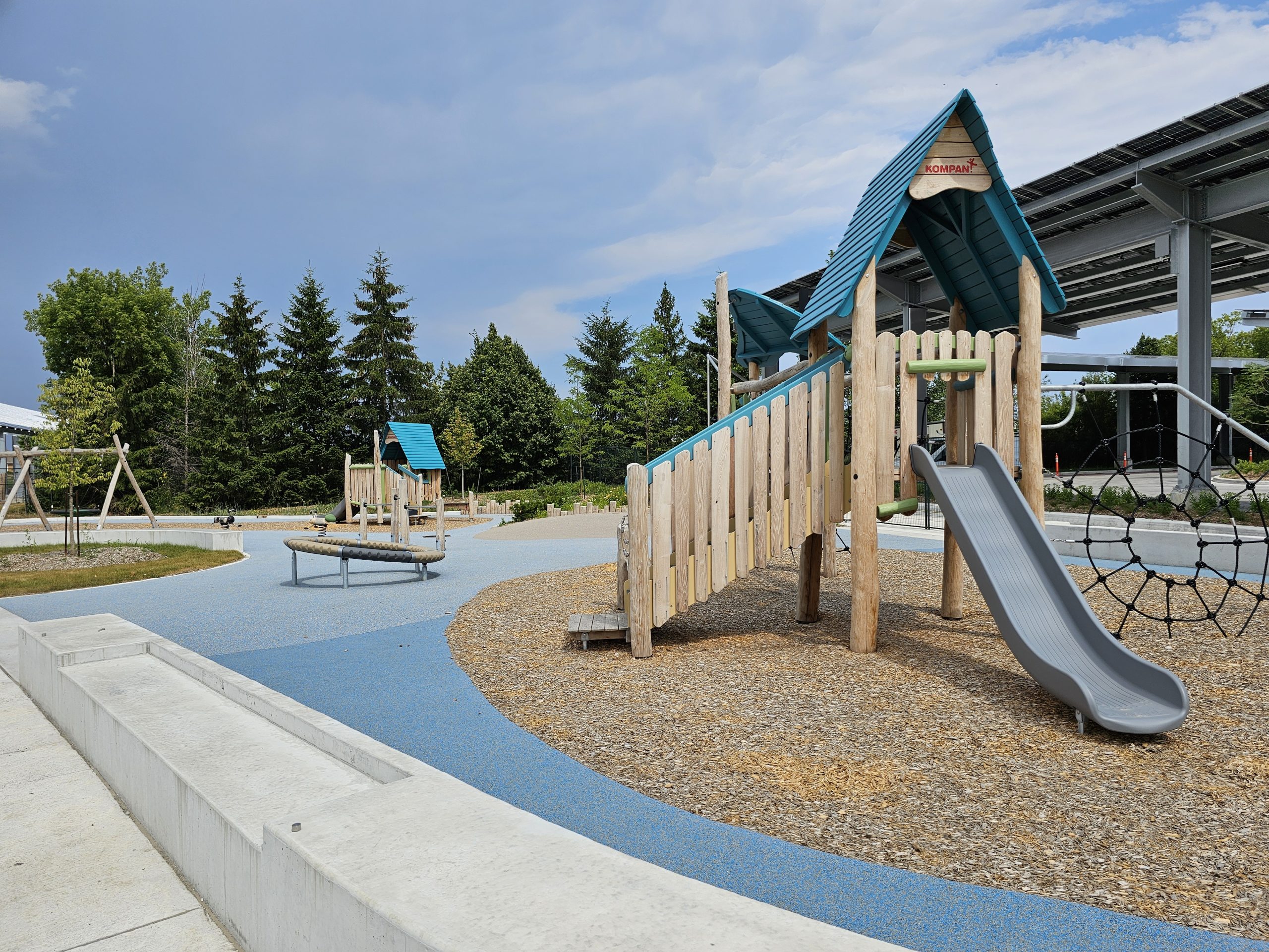 A playground with a wooden climbing structure and slide, surrounded by wood chips and blue rubber flooring. Additional play equipment like swings and a seesaw are visible, with trees and a clear sky in the background.