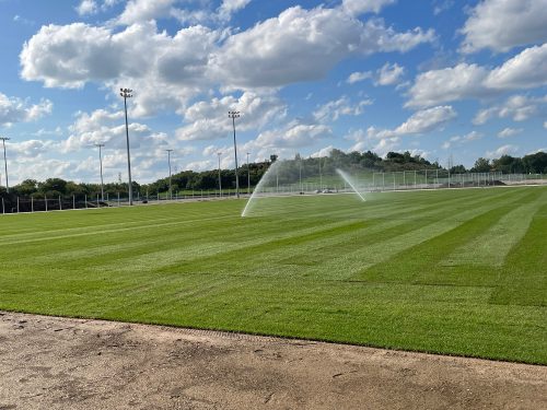 The bubbled training site under construction, showing turf installation and surface irrigation in progress. Two sprinklers are spraying water near the centre of a large grass field.