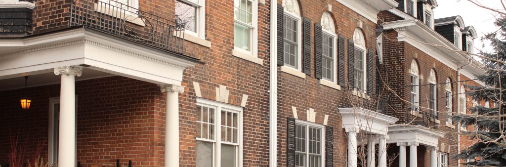 Angled view of two pairs of two-storey, semi-detached Georgian Revival style brown brick buildings on an overcast winter day. The buildings have matching, mirrored façades that create a consistent row along the streetscape. They feature flat roof lines with round arch dormer windows and projecting cornices painted white, and side entrances with Neo-classical porticos.