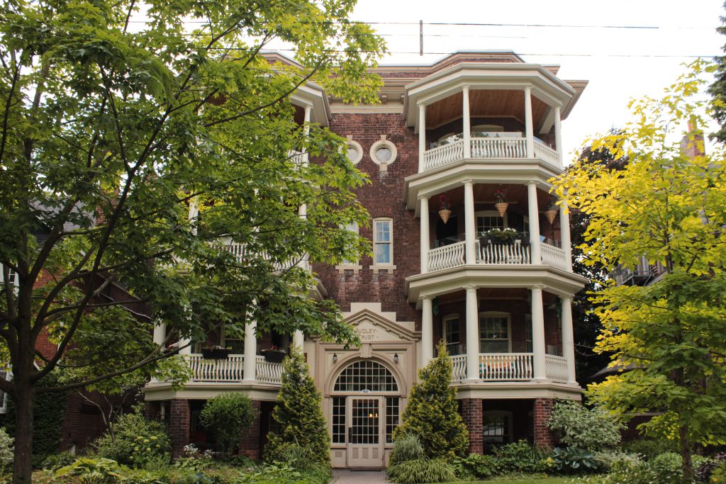 A four-storey, red brick apartment building with three bays and an elaborate central entrance with arched transom window, sidelights, and a relief surround of a triangular pediment. On either side are projecting four-storey, three-sided bay porches with white columns, cornices, and railings. The building is framed by soft landscaping and mature deciduous trees. 