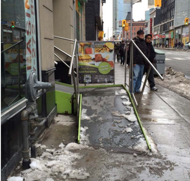 Big black and green ramp with snow - A city sidewalk scene with a temporary ramp installed at a storefront entrance during winter. The ramp is bulky, poorly maintained, covered in snow and ice, and its placement blocks most of the sidewalk. The image illustrates how the ramp creates hazards for pedestrian movement, reducing the clearway and interfering with safe, unobstructed passage