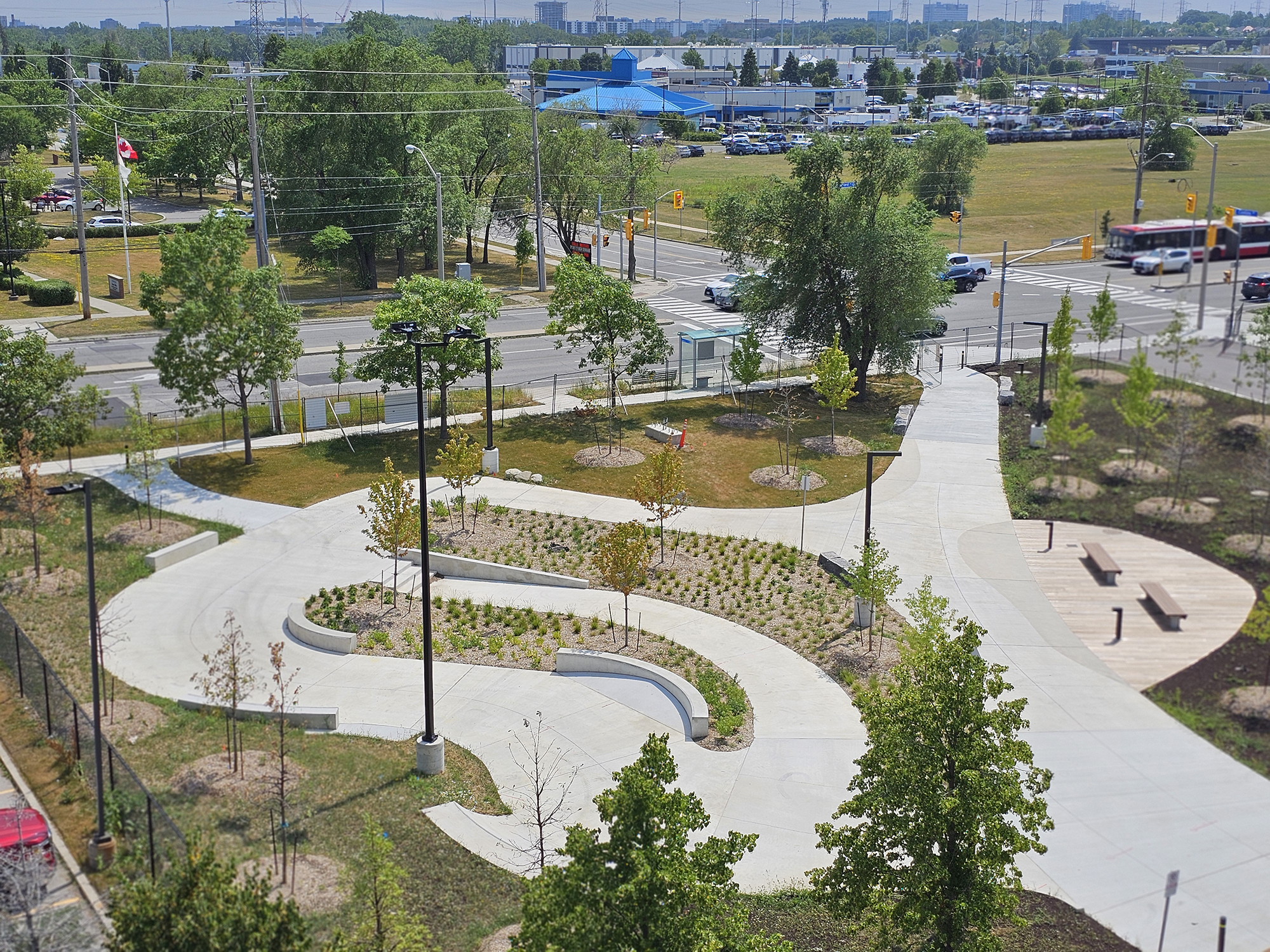 An aerial view of an urban park with winding concrete paths, landscaped areas with young trees and benches, streetlights, and nearby roads with vehicles. Buildings are visible in the distance.