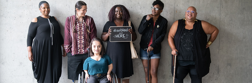 Diverse group of people standing against a concrete wall. One holds a sign reading "Disabled and Here."