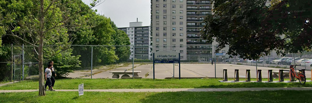 A view of J.A. Leslie Tot Lot from Midland Avenue, facing east showing a large concrete pad with a tall fence around it and a tall apartment building in the background.