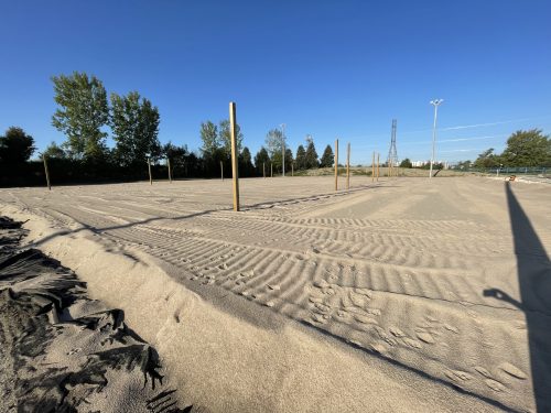 The volleyball courts under construction, showing an even layer of sand with visible tread marks. Several wooden posts are arranged in two rows. Light poles, trees and site fencing are visible in the background.