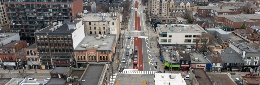 Aerial view of Bathurst Street, looking south from the intersection at Queen Street West, showing a red centre-running streetcar lane with diamond pavement markings. A red TTC streetcar and several vehicles are visible. The street is lined with a mix of brick and modern buildings, and pedestrians are walking along the sidewalks.