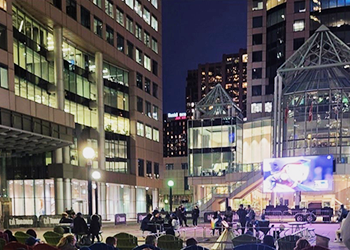 People sitting in an outdoor courtyard watching a baseball ball game at night on a big screen.