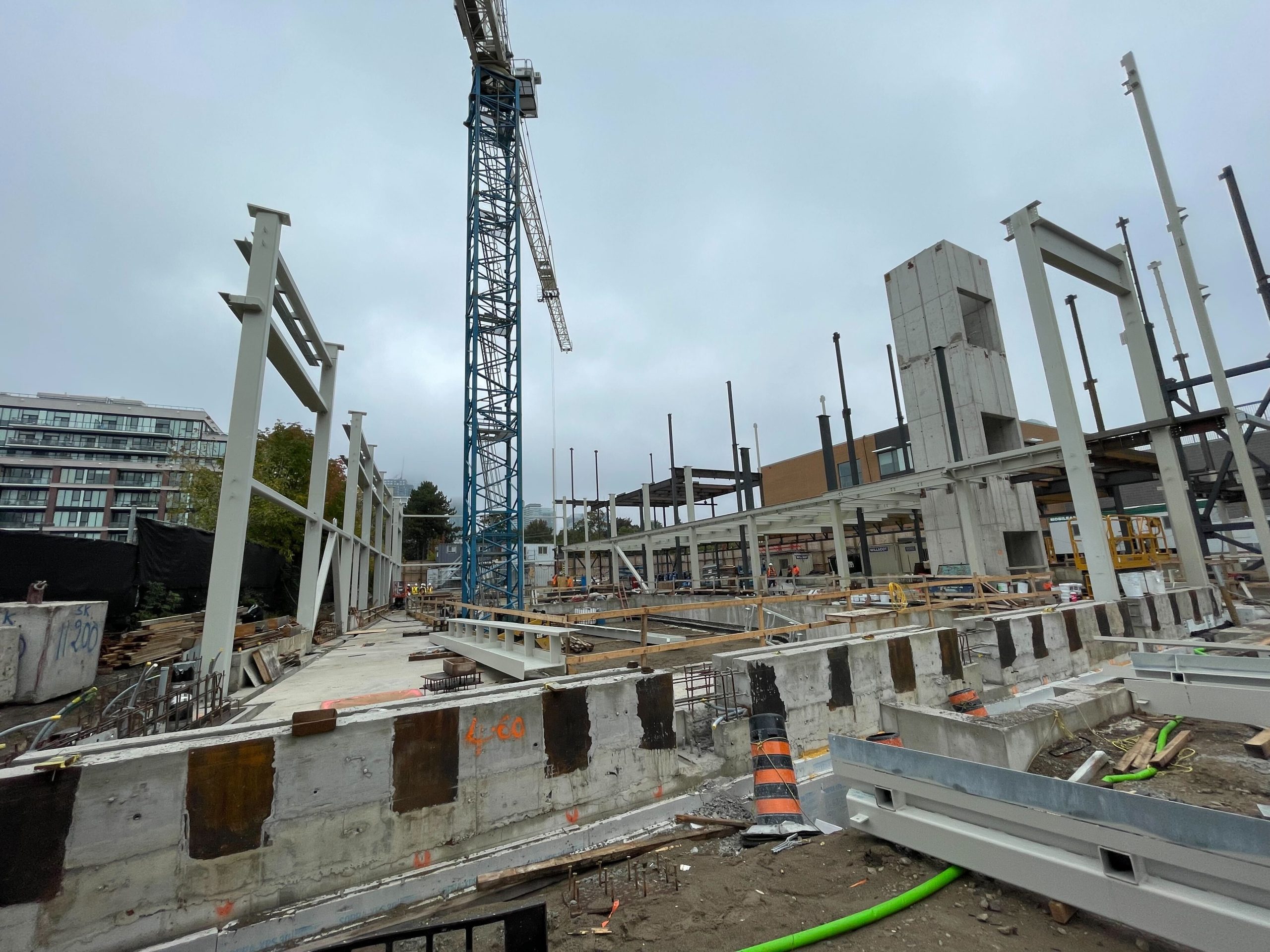 The construction site for the new community centre on an overcast day, showing the installation of steel framing in progress atop a concrete base. A tall crane stands at the centre of the site, surrounded by scattered construction materials and tools. Construction workers are visible in the background.