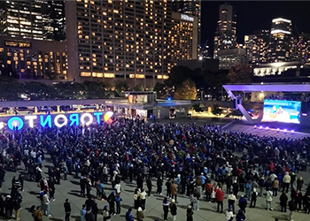 A large crowd of people gathered in a public square at night watching a large screen. A glowing sign in the background reads TORONTO.