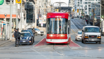 A red streetcar travelling in a priority streetcar lane on Bathurst Street, surrounded by cars and a person cycling near an intersection. Urban buildings and traffic signals are visible in the background.