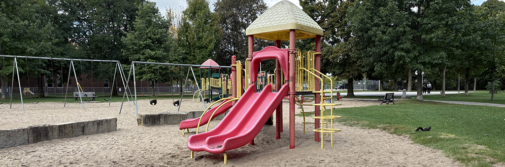A playground with a sandy surface featuring a red and yellow play structure with slides, a climbing ladder, and a small roof. Swings and other equipment are visible in the background, surrounded by grass, trees, and benches under a partly cloudy sky.