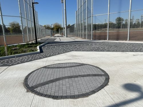 The baseball hub under construction, focusing on the paved walkway between baseball diamonds. The walkway includes a circular design made of light and dark grey bricks arranged to depict a baseball, as well as a patterned border made of grey pavers. Tall light poles and chain-link fences surround the diamonds on either side of the walkway.