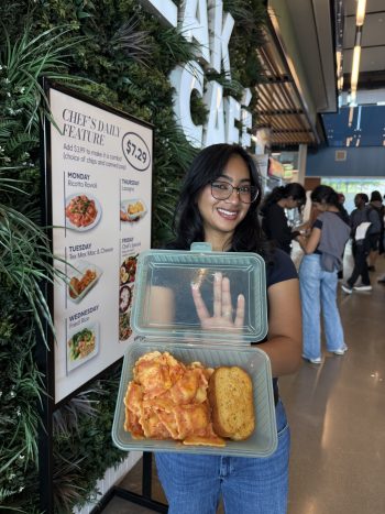 A young woman holding a reusable container full of ravioli and a piece of toast with a sign behind her that reads chef's daily feature.