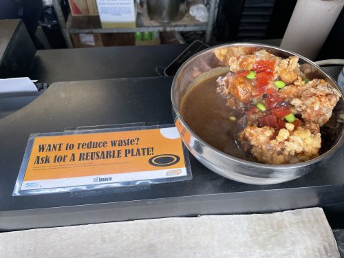 A sign reading want to reduce waste? Ask for a reusable plate! with a reusable bowl beside it full of a dish with sauce, beans, tomatoes and other food.