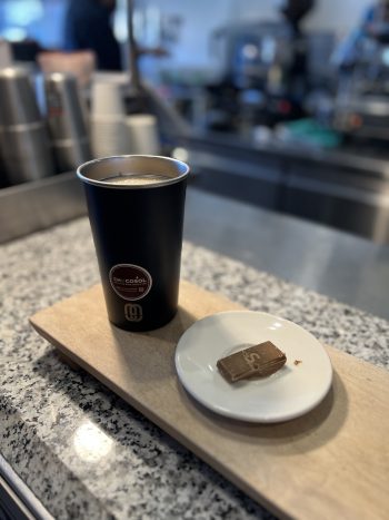 A reusable cup full of a hot beverage and a small plate with a piece of chocolate on a wooden board in the Chocosol cafe and chocolate bar.