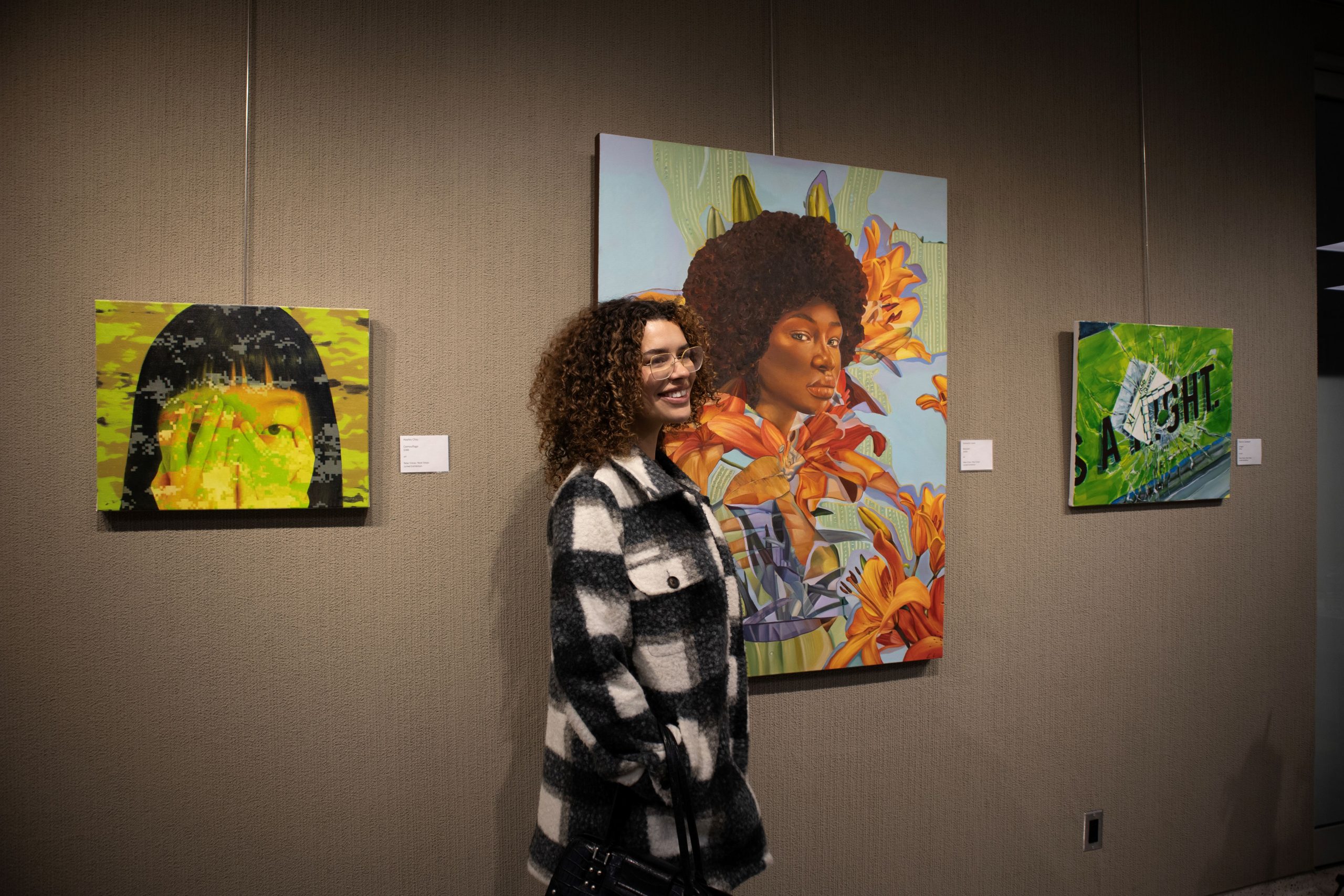An artist stands in front of three artworks displayed in a gallery. The artist is wearing a black and white checkered coat and has curly hair. The artwork on the left features a stylized portrait with green tones and an abstract background. The central piece is a vibrant portrait of a person with an afro hairstyle surrounded by orange flowers. The artwork on the right has green hues with abstract elements.