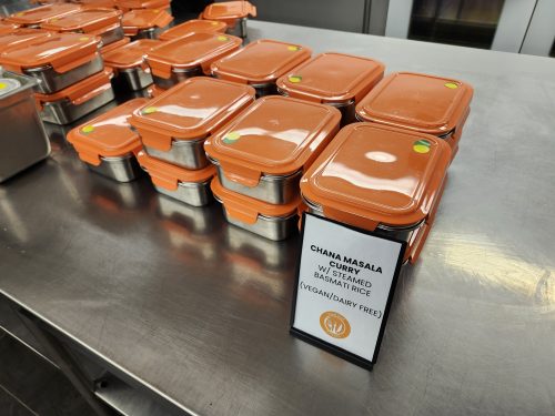 A stack of reusable containers with orange lids with assign reading chana masala curry with steamed basmati rice and vegan/dairy free.