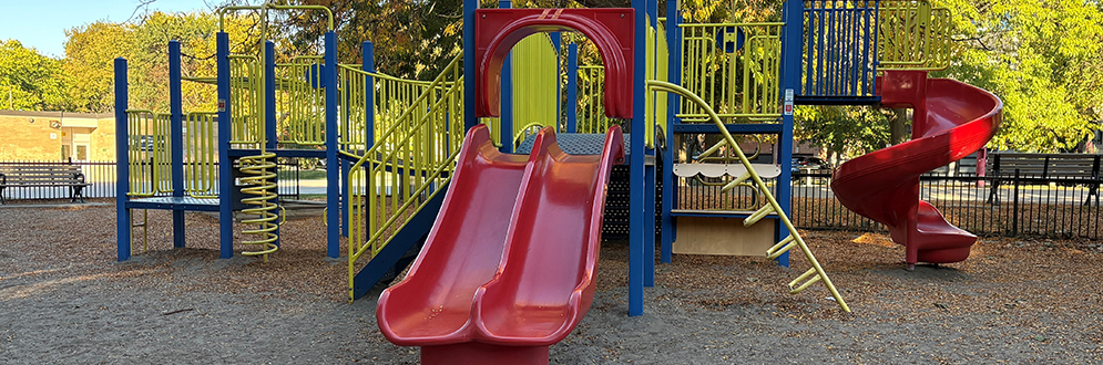 A colourful children's playground with multiple slides, including a double red slide and a spiral red slide, surrounded by blue and yellow climbing structures. The ground is covered in wood chips.
