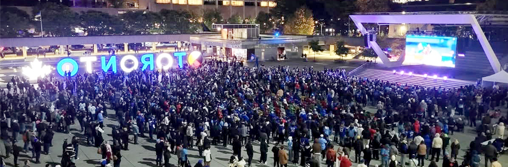 A large crowd of people gathered in a public square at night watching a large screen. A glowing sign in the background reads TORONTO.