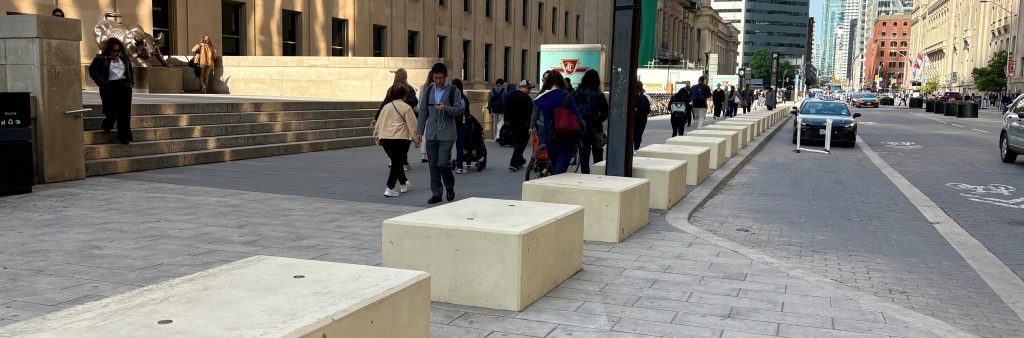 Pedestrians walking along the front plaza of Union Station in Toronto with large rectangular concrete barriers that line the sidewalk beside the street.