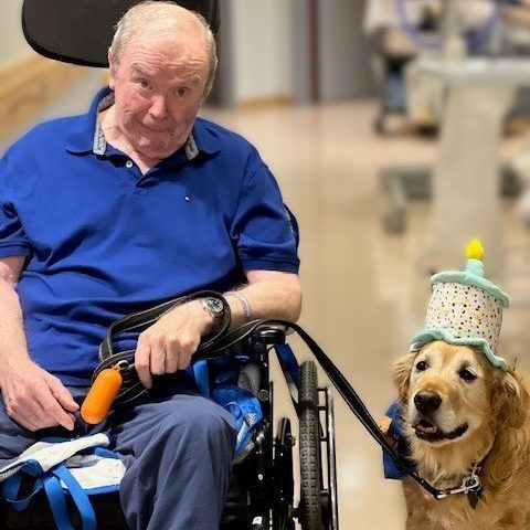A resident wearing a blue golf shirt sits in a wheelchair holding the leash of Bourbon the therapy dog