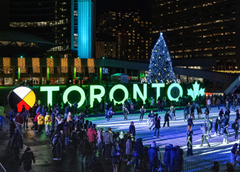 Public Square (Nathan Phillips Sqaure) in front of Toronto City Hall lit up at night with festive lights, the TORONTO sign illuminated in green and lots of people ice skating.