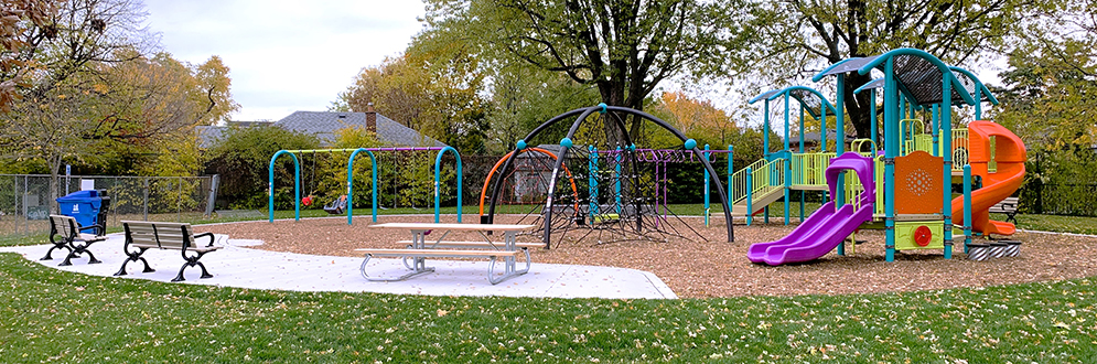 The new Hillside Park playground looking east. The new walkway and seating area are in the front left of the image with the play equipment in the background and to the right. The playground is surrounded by open lawn with mature trees with residences in the background.