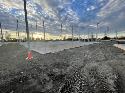 Construction site for new pickleball courts, showing a leveled concrete surface with vertical metal fence posts installed. The ground around the courts is bare soil with visible tire tracks, and orange safety cones are placed near the posts.