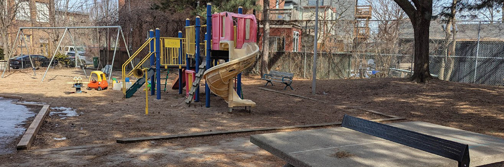 Colourful outdoor playground with slides, climbing steps and platforms. There is a swingset and other play items in the background and a concrete table tennis table in the foreground.