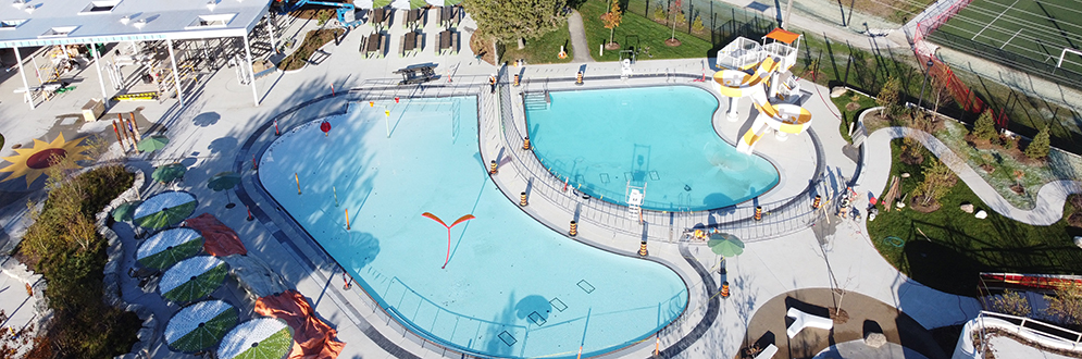 Aerial view of a large outdoor aquatic facility featuring two connected pools with curved edges. The pools are surrounded by concrete walkways, lounge chairs, and green umbrellas. A white shade canopy covers part of the seating area on the left. In the background, a parking lot, sports fields, and autumn-colored trees are visible.