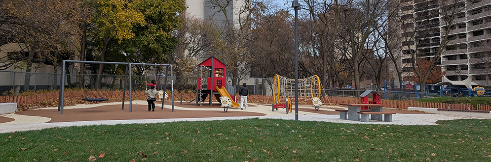 A playground that features swings, a red play structure with a slide, and climbing equipment. There is a green grassy area in the foreground and a paved walkway around the play area.