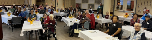 A large group of people is seated at multiple rectangular tables covered with white tablecloths in an auditorium. Each table has a small centrepiece with yellow flowers in a vase. The attendees are facing toward the front, suggesting a presentation or event is taking place.