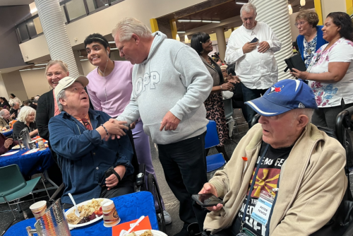 A Thanksgiving celebration at Humber College with people gathered around tables covered in blue tablecloths. The Premier of Ontario, Doug Ford, shakes hands with a seated individual who has a plate of food and a drink in front of them. Another person in a wheelchair wearing a beige sweater and a blue baseball cap is holding a smartphone. The background shows more attendees standing and chatting, and plates of food, cups, and event materials are visible on the tables.
