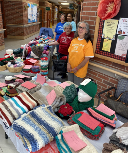 A display table in a hallway is filled with colourful hand-knitted items, including scarves, hats, dishcloths, and other accessories. Several baskets hold smaller knitted pieces, and mannequin heads showcase hats and scarves. Pink price tags are placed on many of the items. Behind the table, three individuals wearing bright T-shirts with the words “Knotty Knitters” stand near the display. A bulletin board with notices and decorations is visible on the wall in the background.