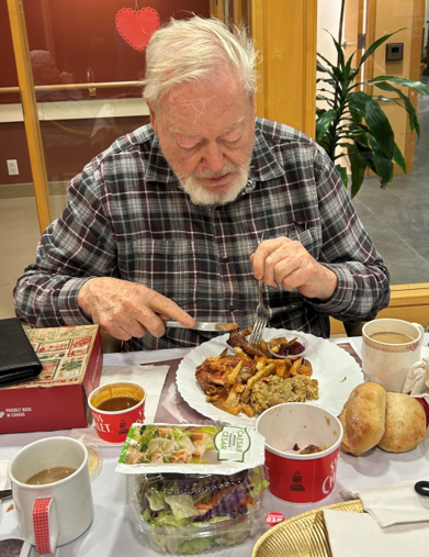 A person seated at a dining table is eating a chicken dinner. The table is covered with a white tablecloth, and surrounding the plate are a salad container, two bread rolls, a cup of soup, two mugs, and a red takeout box.