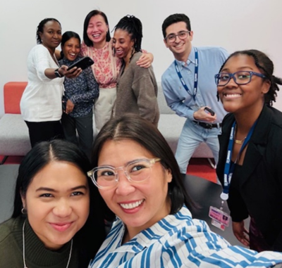 A group of eight people gathered indoors, posing for a casual photo. Some are standing while others lean in close, and one person is holding a smartphone as if taking a selfie. The background includes a light-colored wall and a red-and-gray cushioned bench.