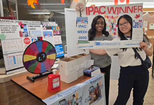 Two people stand behind a display table for IPAC Week 2025 at Lakeshore Lodge. They are holding a large rectangular frame with the words “IPAC WINNER,” and the centre of the frame has been cut out so their faces can peek through. The table in front of them features educational materials on infection prevention and control, a colourful prize wheel, a red hand sanitizer dispenser, and several boxes. Behind the table are posters with information about hand hygiene and infection control.