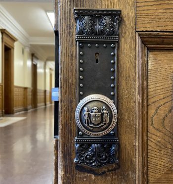 Close-up of an ornate metal door plate and round knob on a wooden door in Old City Hall. The plate features decorative scrollwork and a row of rivets, with a keyhole in the center. The knob displays an embossed crest with detailed figures and shield design. 