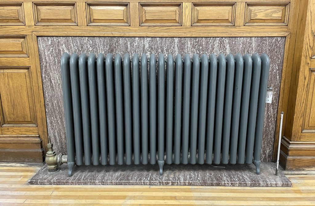 Close-up of a vintage cast-iron radiator with vertical columns painted gray, set against a polished marble panel and surrounded by wood-paneled walls. Brass valve hardware is visible on the left side, and the radiator sits on a marble base above a hardwood floor.