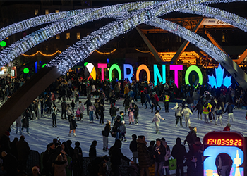 A busy outdoor skating rink at night lit up by white twinkly lights overhead and large letters lit in bright colours that spells TORONTO.