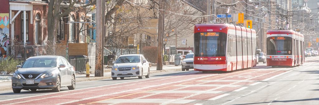 Two TTC streetcars in red-painted transit priority lanes with cars in an adjacent lane on a city street lined with houses, mid-rise buildings and utility poles.