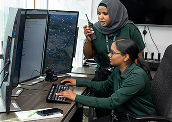 A crisis support worker talking into a walkie-talkie and the other sitting at a desk typing as they look at computer screens.