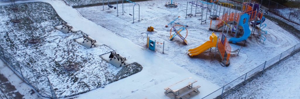 Aerial view of the new Indian Line Park playground looking southwest with a new walkway continuing to the shade shelter and benches to the left and the playground with wood fiber surfacing to the right. The playground is surrounded by open lawn and mature trees with tennis courts further in the background.